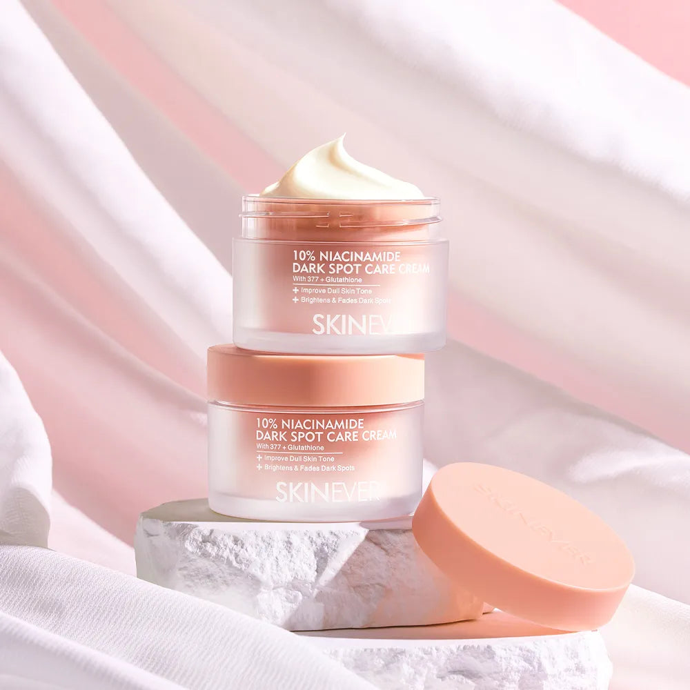 Two SKINEVER 10% Niacinamide Dark Spot Care Cream jars stacked on a white stone slab, with one jar open to show a pale cream product and the other jar with its peach-colored lid resting beside it. The background consists of draped white and light pink fabric.